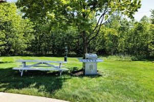 Una mesa de picnic y una parrilla en un parque con césped y árboles al fondo.