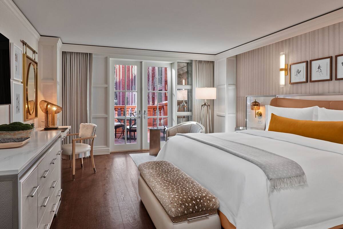 A hotel room with a large bed, a desk with a lamp, a chair, and a bench. There is a view of the outside through a glass door.