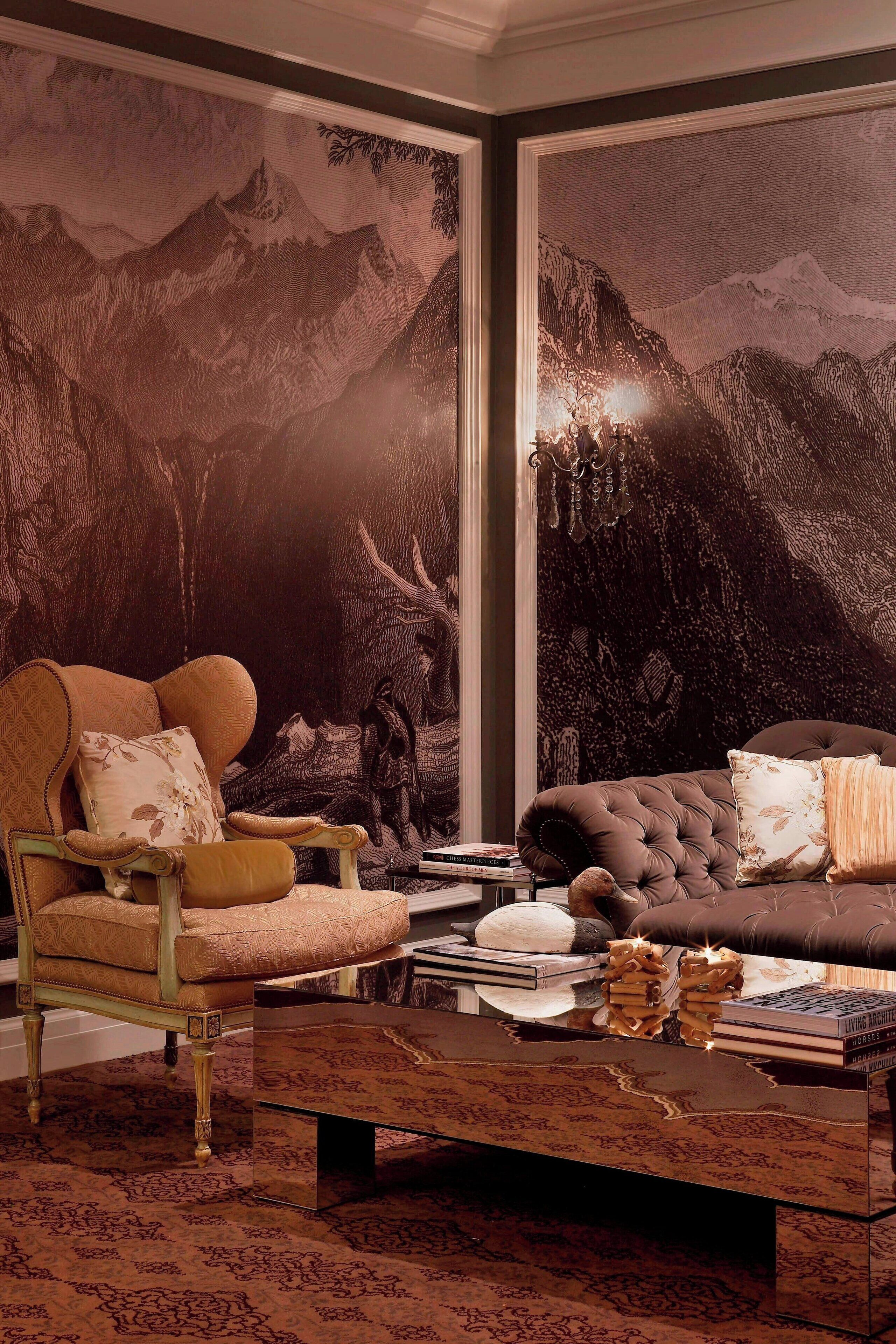 A room with a large wall mural of mountains, a plush sofa, a vintage armchair, and a coffee table with books and decorative items.