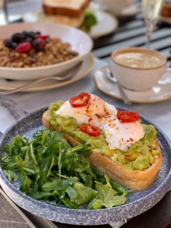 A plate with a sandwich topped with avocado, poached egg, and red chili, accompanied by a side of greens.