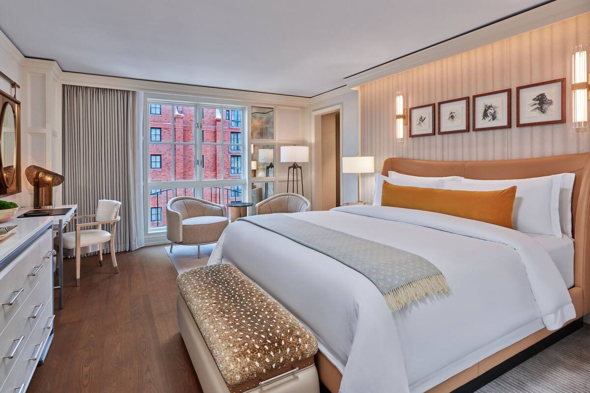 A hotel room with a large bed, a desk with a lamp, a chair, a bench, and a view of a red brick building through a large window.