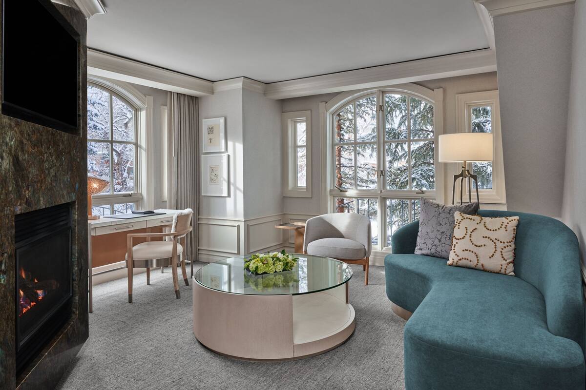 A modern living room with a glass coffee table, a teal sofa, a fireplace, and large windows with a view of snow-covered trees.