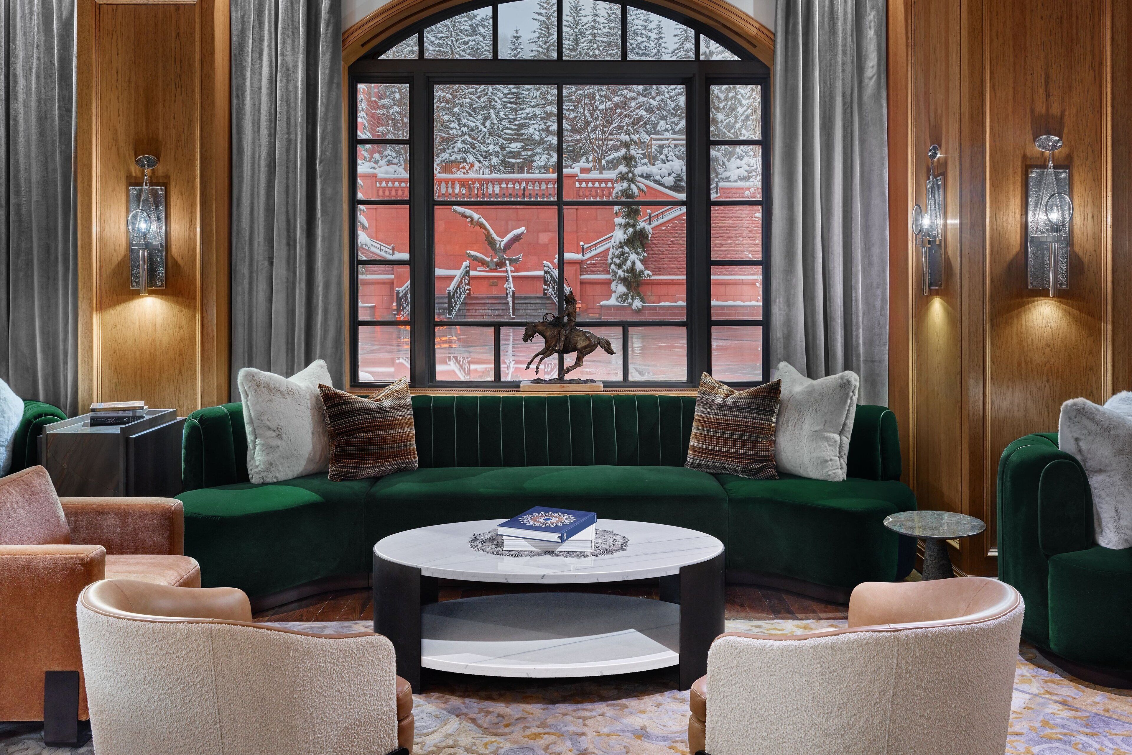 Un salón acogedor con un sofá verde, sillones beiges y una mesa de centro redonda. En el fondo, se ve una ventana con vistas a un paisaje nevado y una estatua de un caballo.