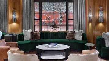 Un salón acogedor con un sofá verde, sillones beiges y una mesa de centro redonda. En el fondo, se ve una ventana con vistas a un paisaje nevado y una estatua de un caballo.