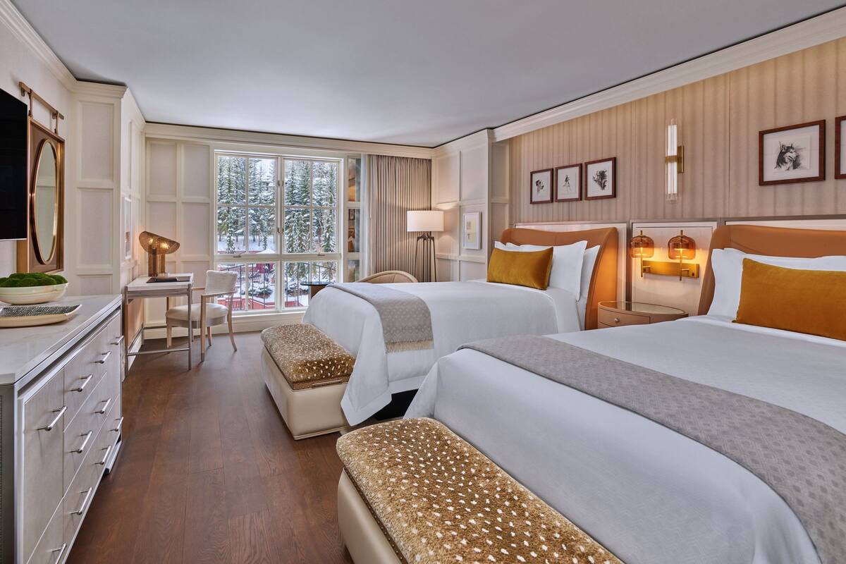 A hotel room with two beds, a desk, a chair, a dresser, and a large window with a view of trees.