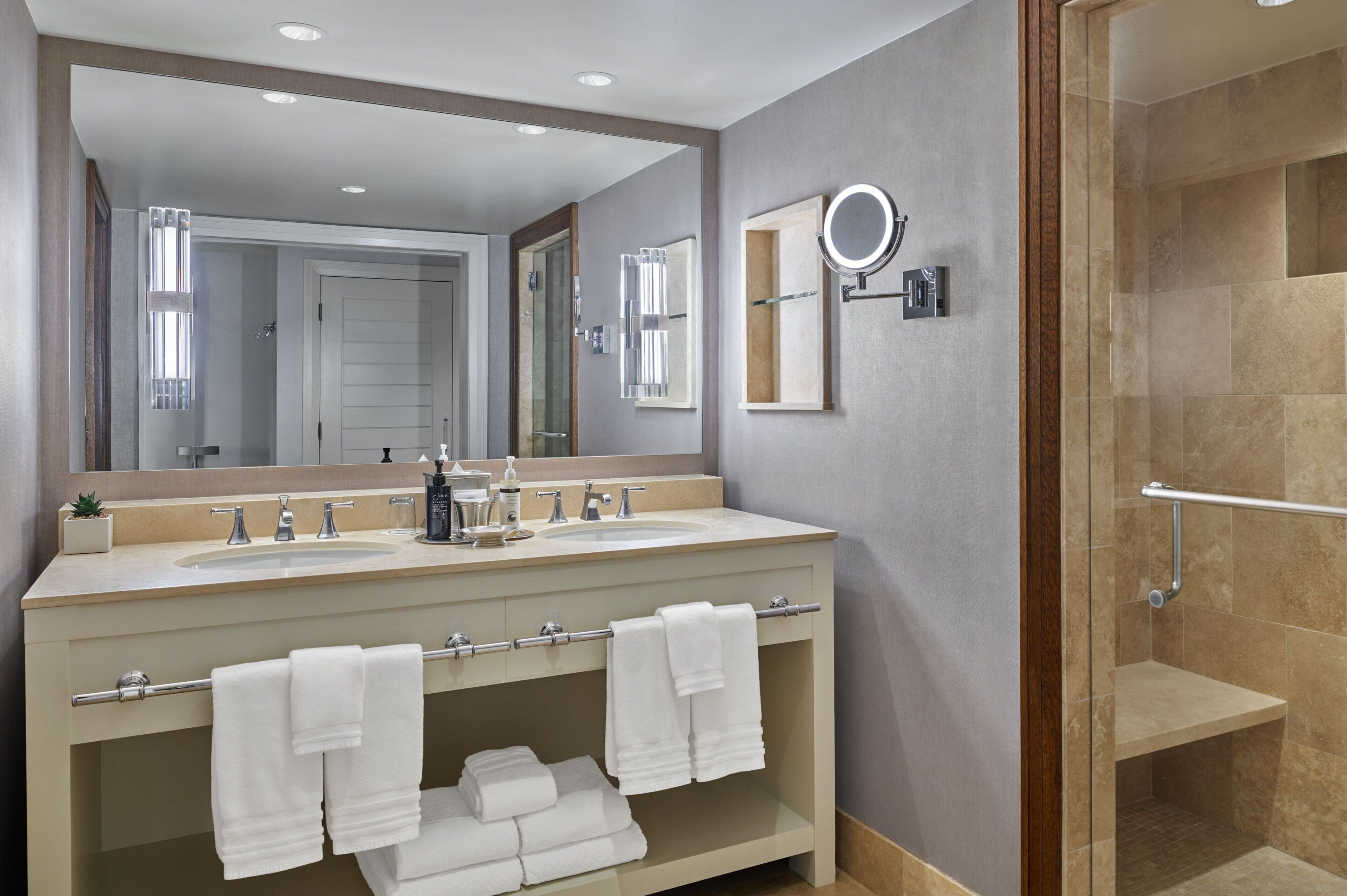 A modern bathroom with two sinks, a large mirror, a handheld mirror, and a glass-enclosed shower.