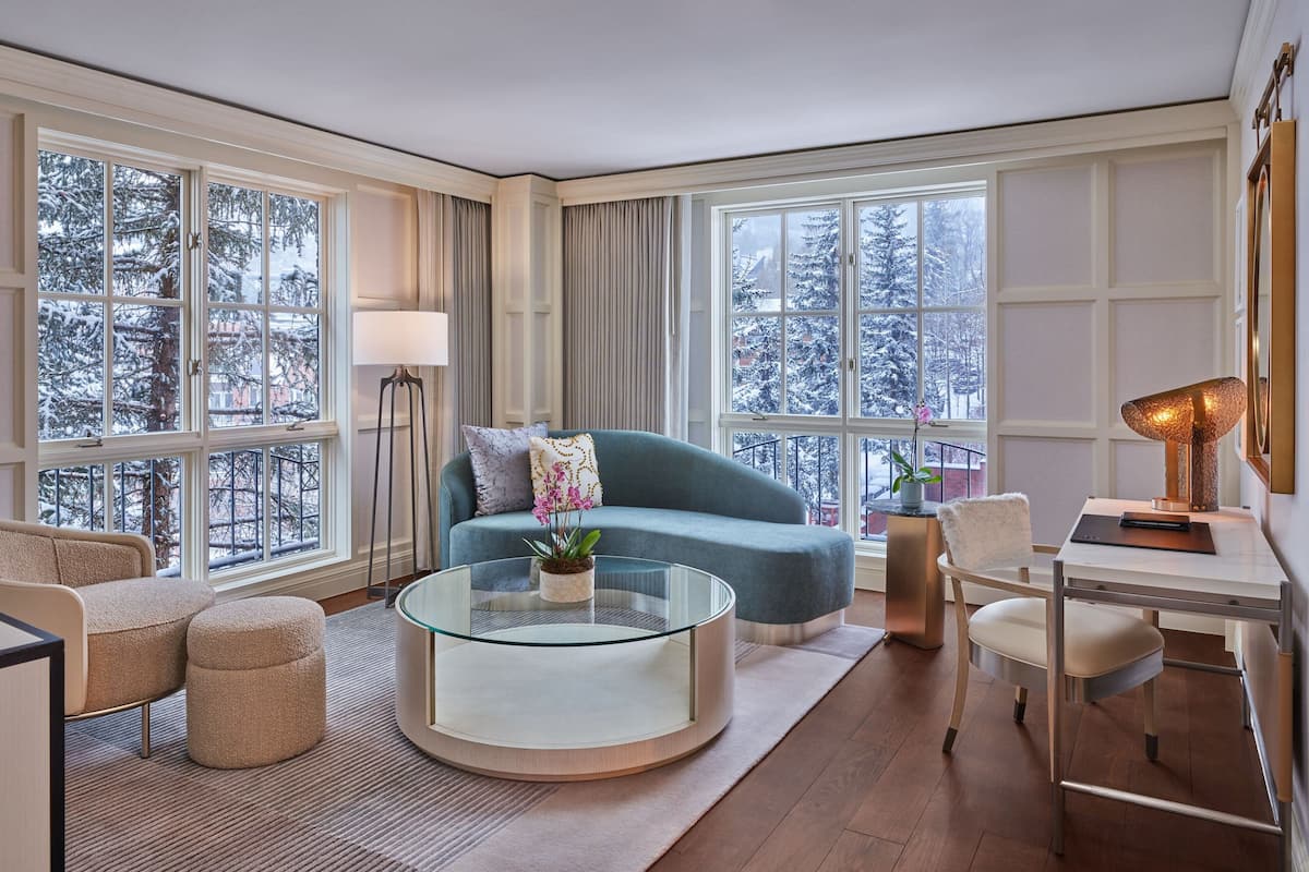 A modern living room with a blue sofa, a glass coffee table, and a desk with a lamp. Large windows offer a view of snow-covered trees.