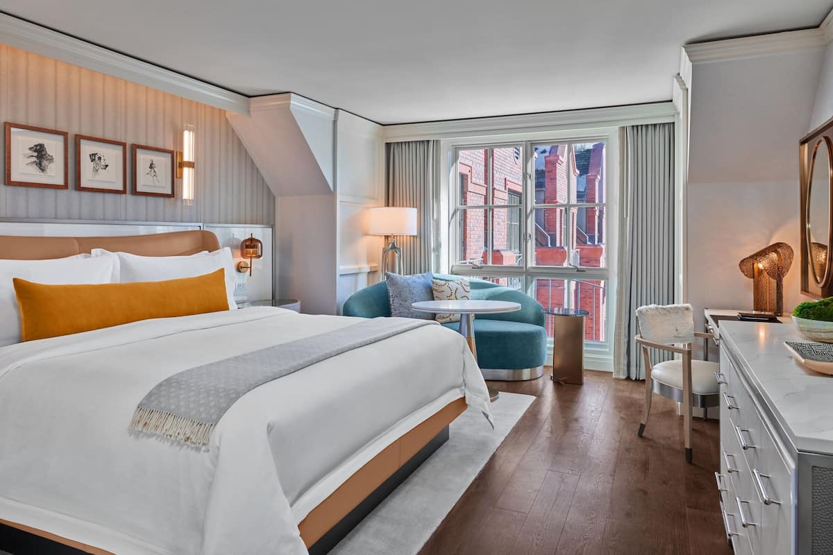 A hotel room with a large bed, a desk, a chair, a small round table, and a view of the city through a window.