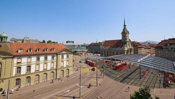 Ein historisches Gebäude mit einer grünen Kuppel und einem Glockenturm, umgeben von einer Straßenbahnhaltestelle und einem modernen Glasbau.