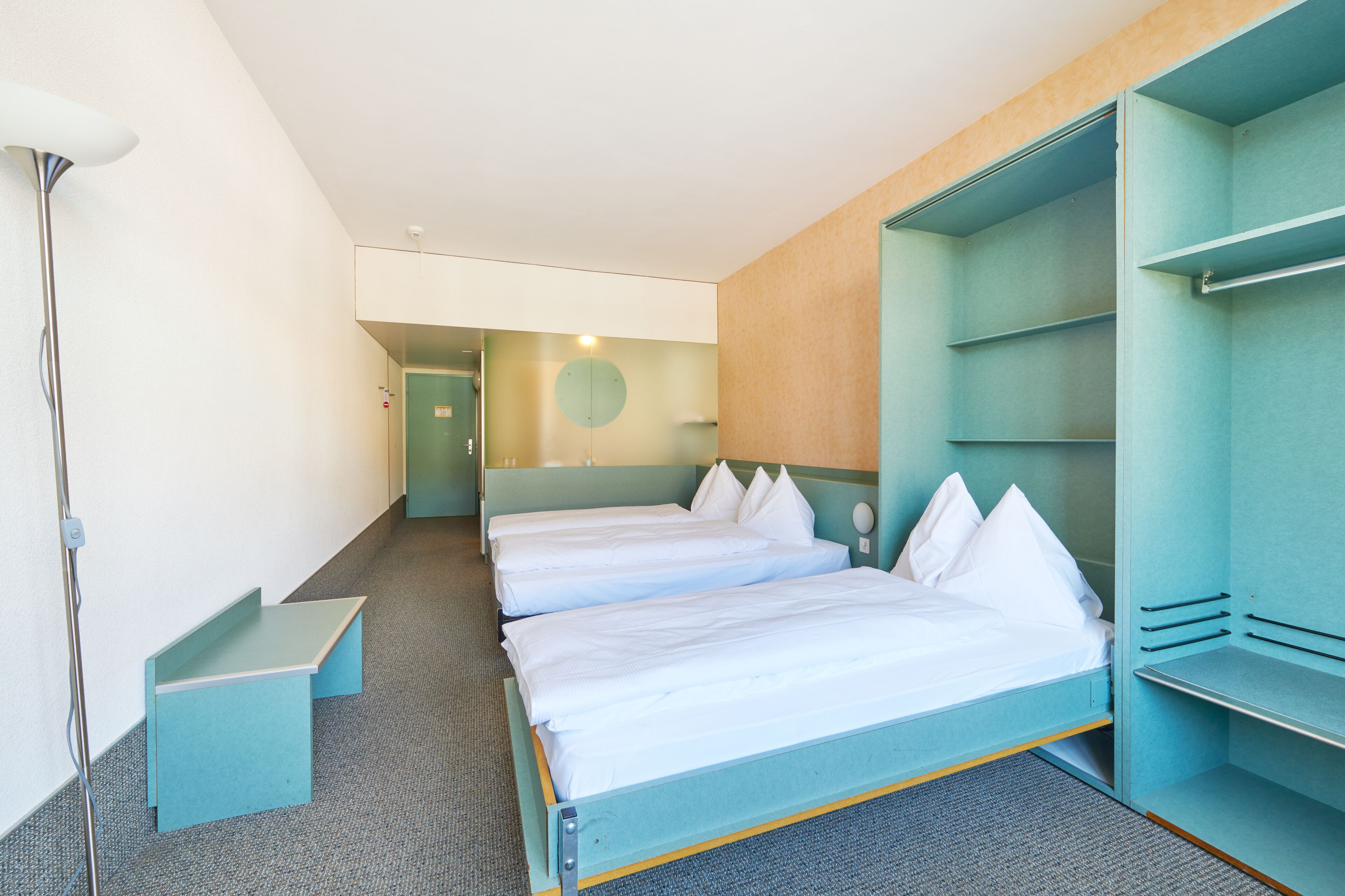 A hotel room with two beds, a desk, a wardrobe, and a lamp.