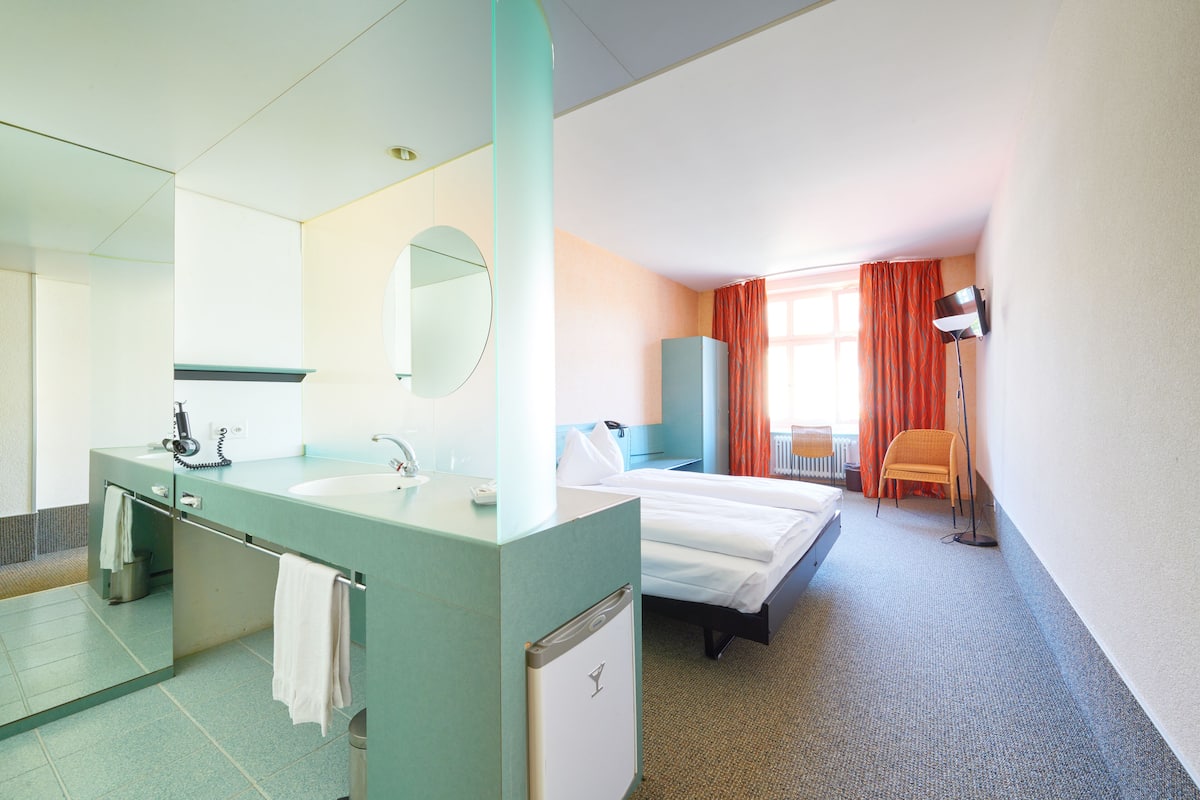 A hotel room with a large mirror, a sink, a bed, a chair, and a lamp.