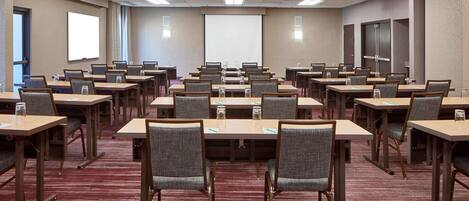 A conference room with multiple tables and chairs, equipped with water glasses and notepads, and a large projection screen on the wall.