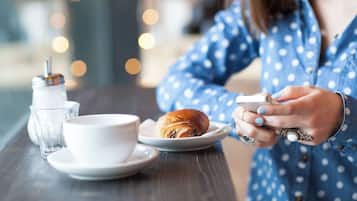 Una persona sosteniendo un teléfono inteligente en una mesa con un croissant, una taza y un vaso de agua.