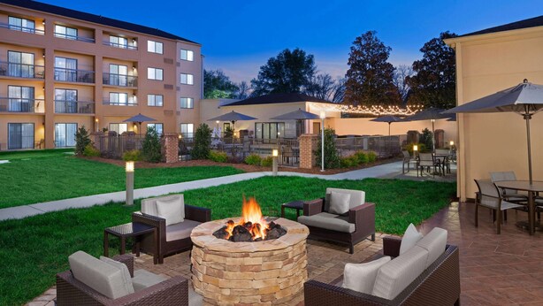 A patio area with a stone fire pit surrounded by wicker furniture, outdoor lighting, and a view of a multi-story building in the background. - Sonesta Select Raleigh Durham Airport Morrisville (Morrisville)