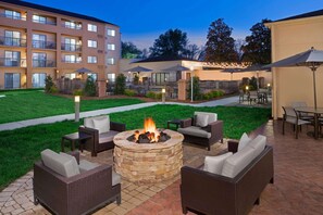 A patio area with a stone fire pit surrounded by wicker furniture, outdoor lighting, and a view of a multi-story building in the background. - Sonesta Select Raleigh Durham Airport Morrisville (Morrisville)