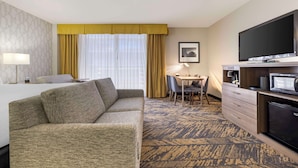 Habitación de hotel con sofá, mesa de comedor con sillas, televisor, refrigerador y alfombra estampada.