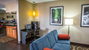 A modern hotel room with a blue sofa, a small desk with a laptop, a coffee machine, and a framed forest picture on the wall.