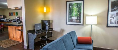 Een moderne hotelkamer met een blauwe bank, een klein bureau met een laptop, een koffieapparaat en een ingelijst boslandschap aan de muur.