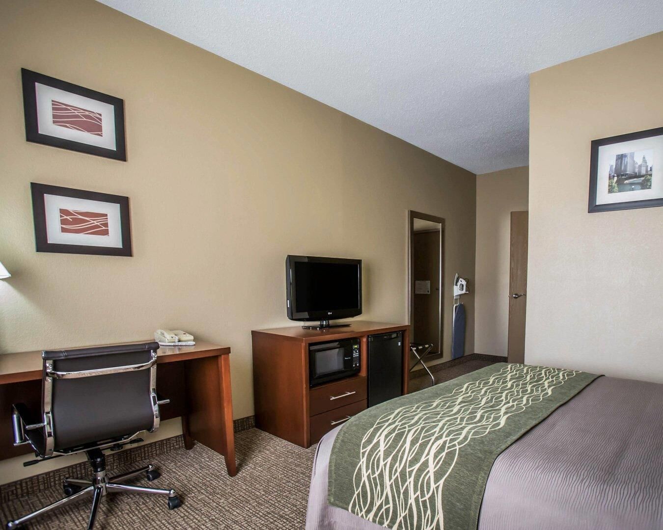 Una habitación de hotel con una cama, un escritorio con una computadora, un televisor y dos cuadros en la pared.