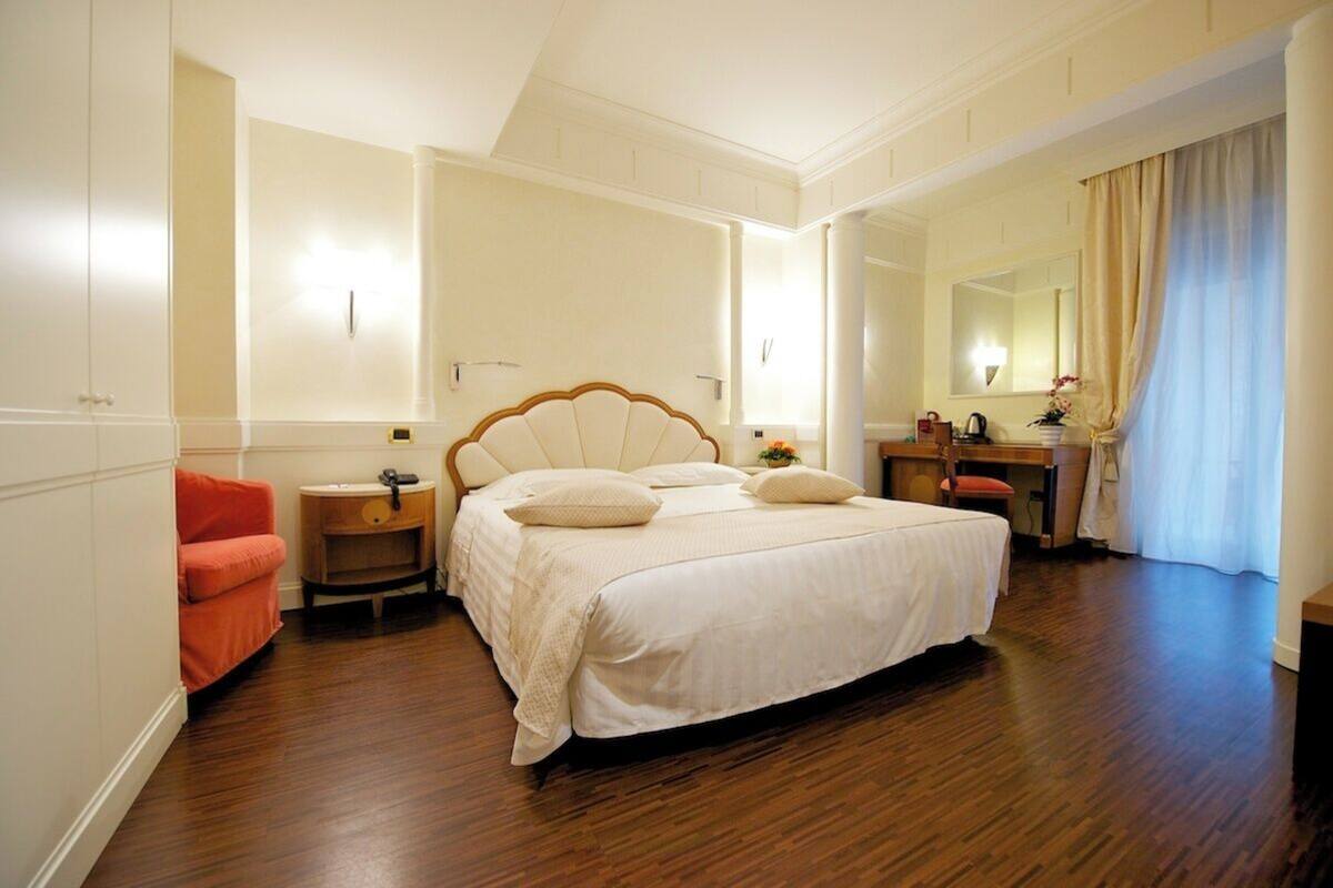 A bedroom with a large bed, a bedside table, a chair, a desk, and a mirror.