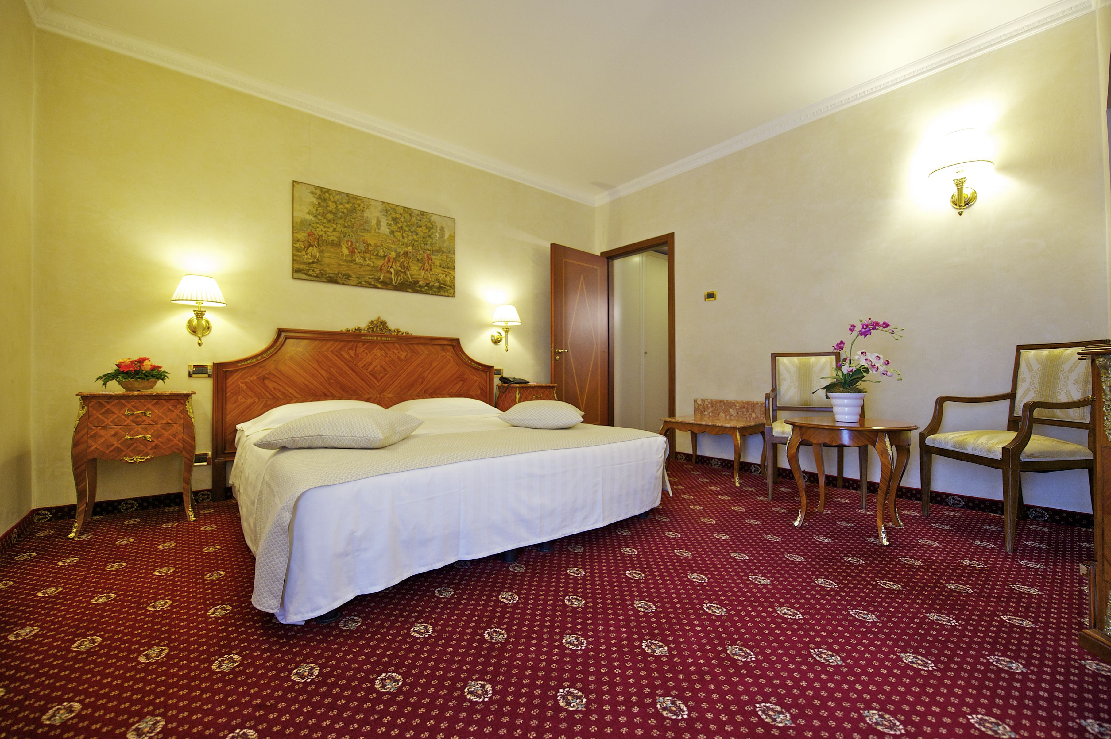 Ein Hotelzimmer mit einem großen Bett, zwei Nachttischen mit Lampen, einem Sessel, einem kleinen Tisch mit Blumentopf und einem Bild an der Wand.