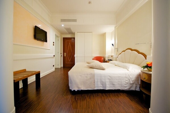 A hotel room with a bed, a bench, a TV mounted on the wall, and a door in the background. - HOTEL STENDHAL (Parma)