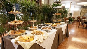A buffet table with various food items and a tiered dessert display in a well-lit room with a tiled floor and potted plants.