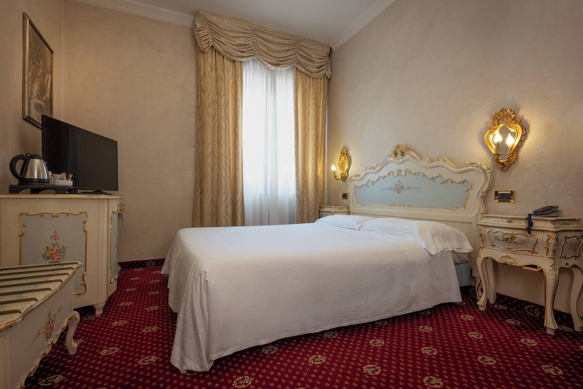 A hotel room with a large bed, a television, a bedside table with a lamp, and a red patterned carpet.