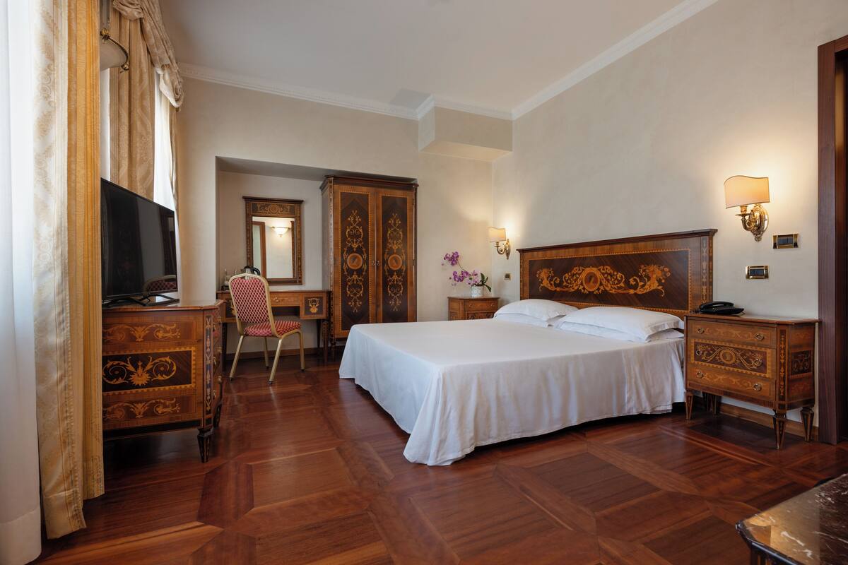 A hotel room with a large bed, wooden furniture, a desk, a television, and a mirror.