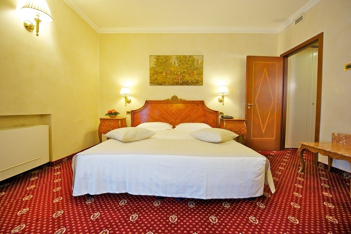 A hotel room with a large bed, two bedside lamps, a wooden headboard, a red patterned carpet, and a painting on the wall.