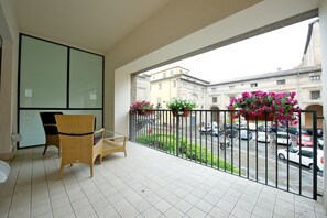 A balcony with wicker chairs, a glass partition, and a view of a street with parked cars and buildings.