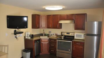 A compact kitchen with wooden cabinets, a built-in microwave, a stainless steel refrigerator, and a flat-screen TV mounted on the wall.