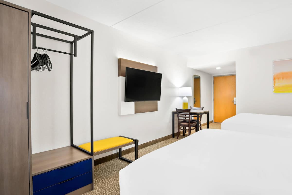 A hotel room with a bed, a desk with a chair, a bench, a wardrobe, and a TV mounted on the wall.