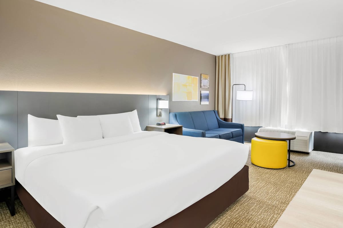 A hotel room with a large bed, a blue armchair, a yellow ottoman, a small round table, and a nightstand with a lamp.
