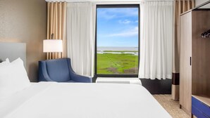 A hotel room with a large bed, a chair, a wardrobe, and a window with a view of a green landscape. - Comfort Inn Gold Coast (Ocean City)