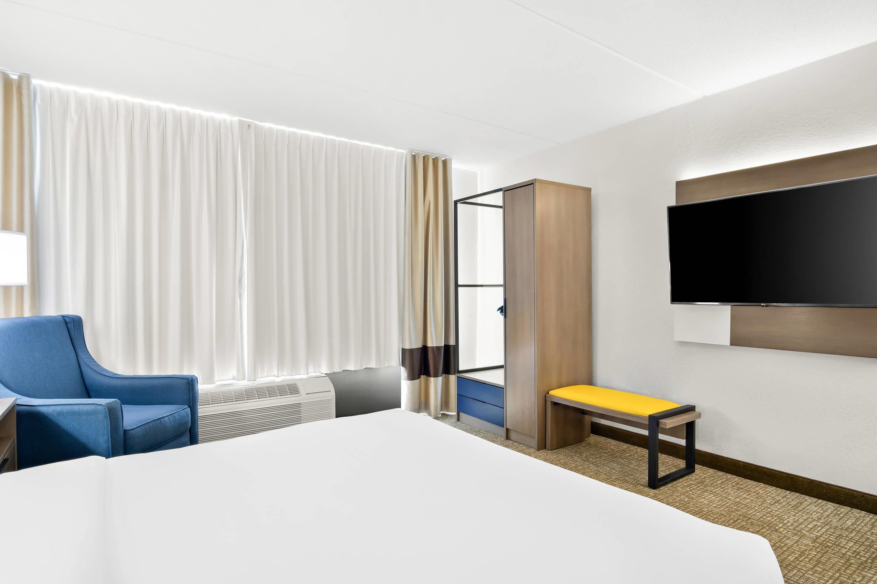 A hotel room with a large bed, a blue armchair, a bench, a TV mounted on the wall, and a window with curtains.