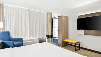 A hotel room with a large bed, a blue armchair, a bench, a TV mounted on the wall, and a window with curtains.