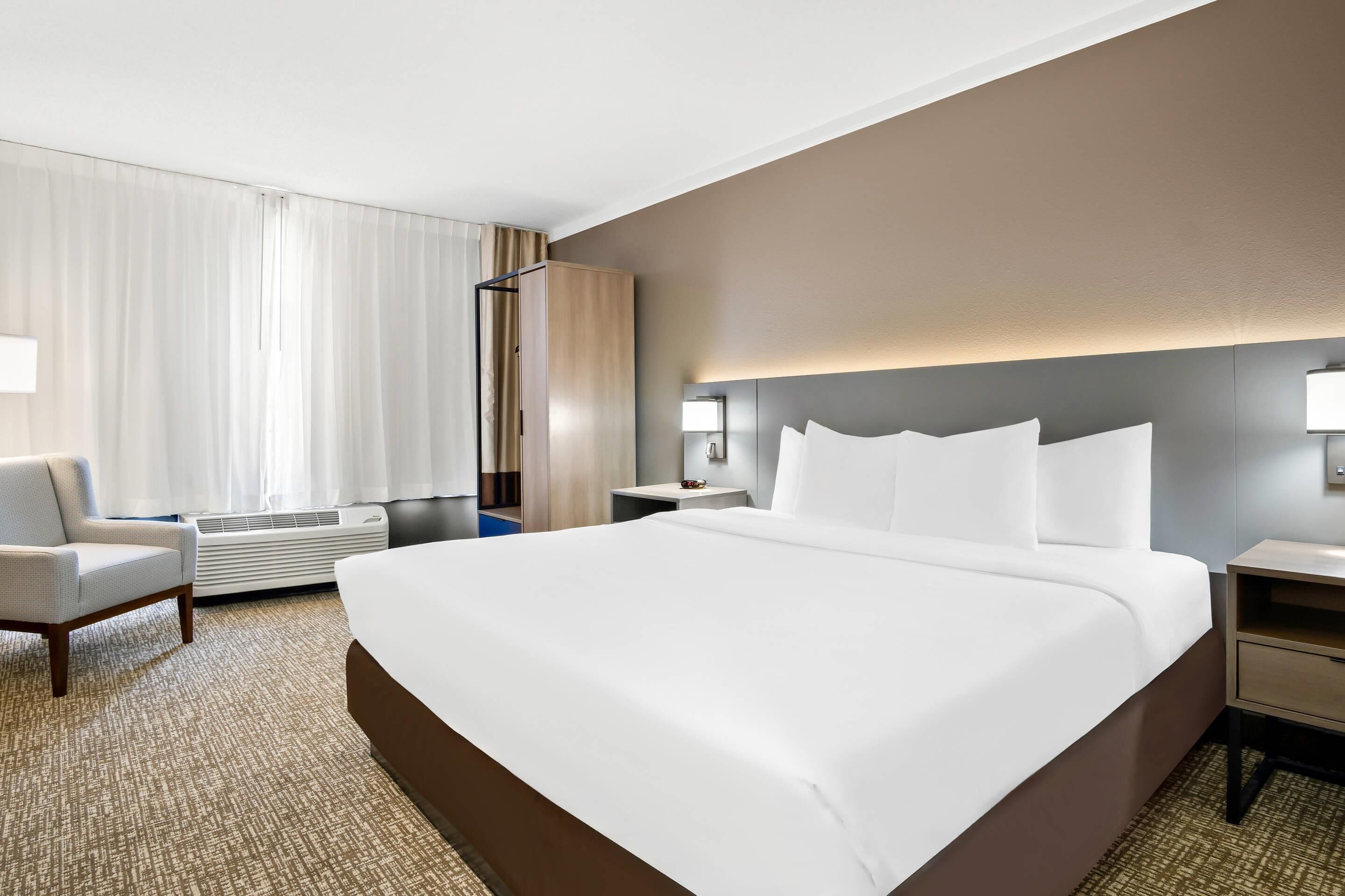 Una habitación de hotel con una cama grande, una silla, una mesita de noche y una lámpara de pared.
