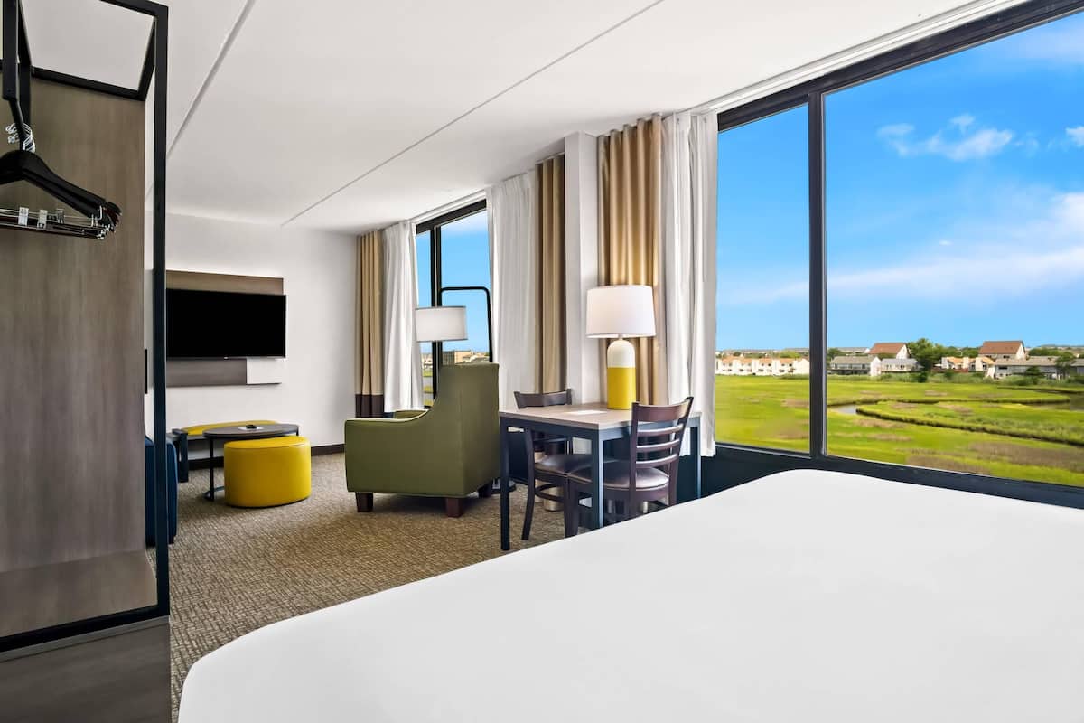 A hotel room with a large bed, a dining area with a table and chairs, a TV mounted on the wall, and a view of a green landscape through large windows.