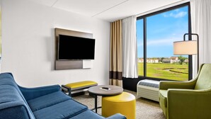 A modern hotel room with a large window, a flat-screen TV, a blue sofa, a yellow ottoman, and a green armchair. - Comfort Inn Gold Coast (Ocean City)