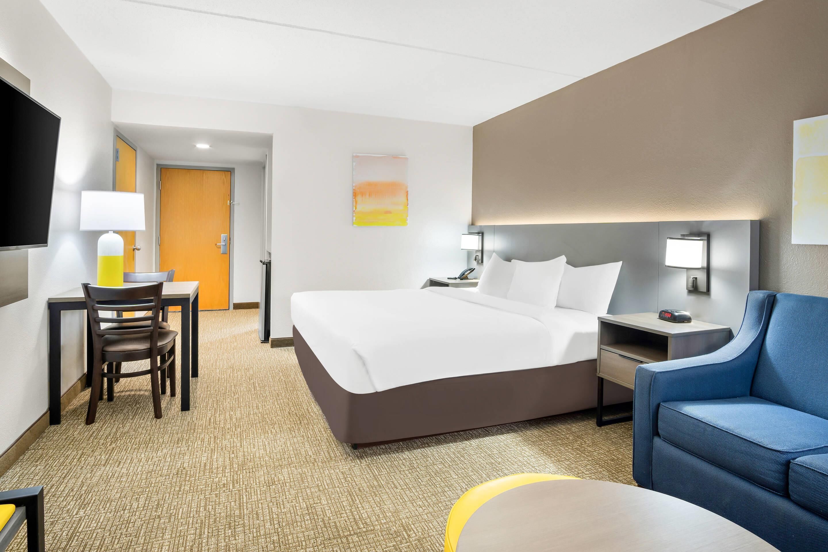 Una habitación de hotel con cama, un escritorio con silla, un sillón azul, un televisor y un cuadro en la pared.