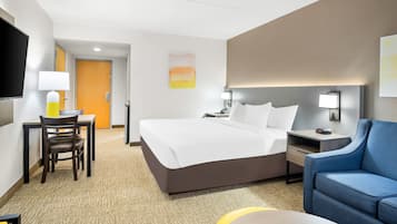 Una habitación de hotel con cama, un escritorio con silla, un sillón azul, un televisor y un cuadro en la pared.