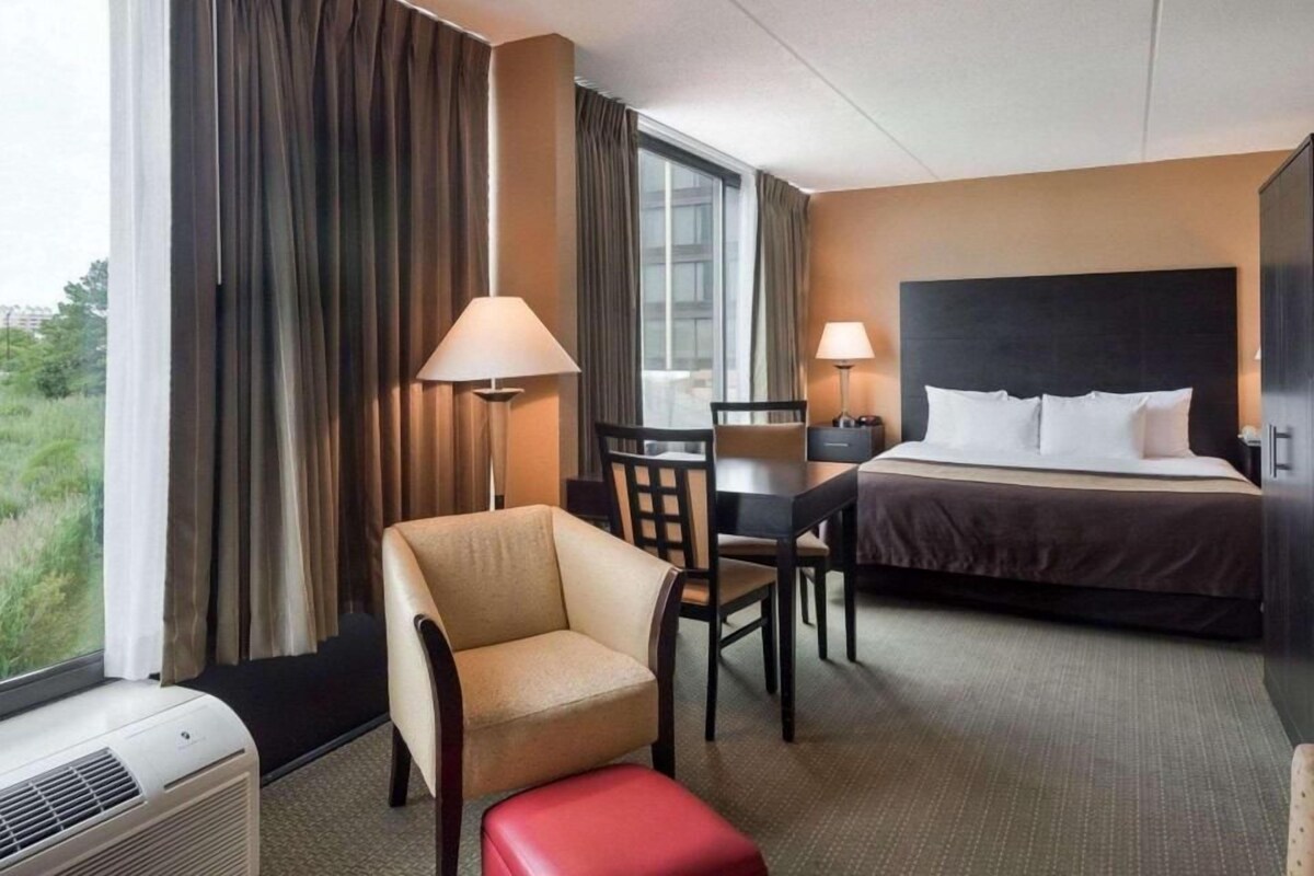 A hotel room with a bed, a chair, a desk, a lamp, and an air conditioning unit.