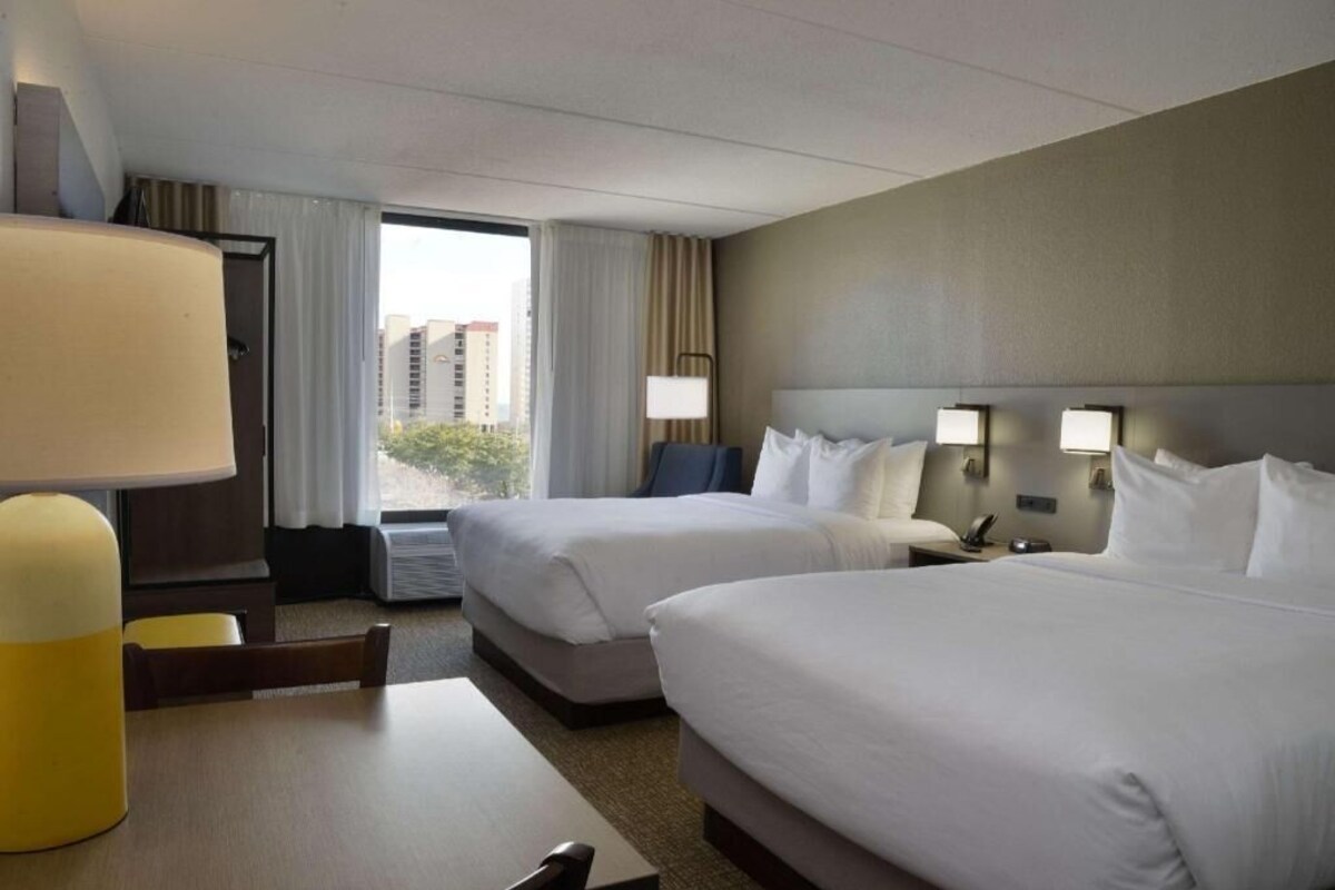 A hotel room with two beds, a desk, a lamp, and a view of buildings through the window.