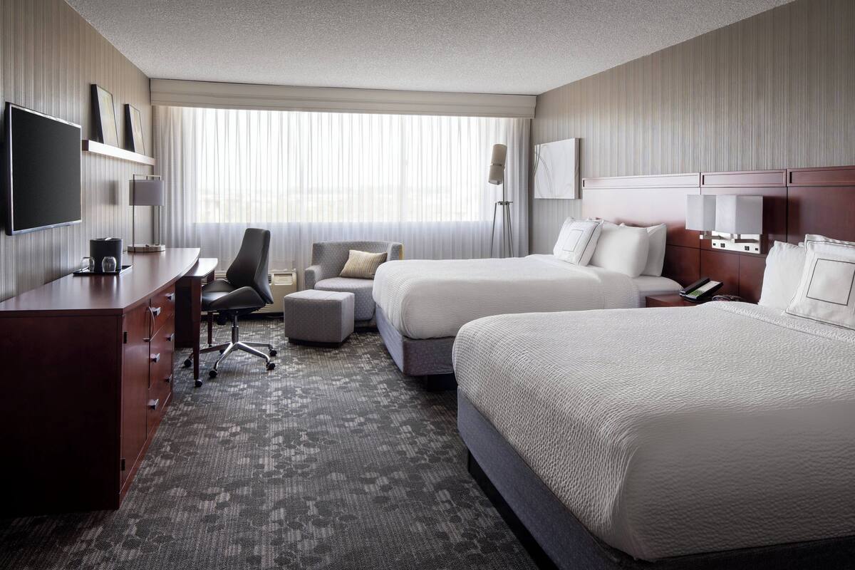 A hotel room with two beds, a desk, a chair, a TV, and a large window with curtains.