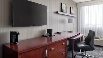A hotel room with a flat-screen TV mounted on the wall, a wooden desk with a coffee maker, a chair, and a small shelf with framed pictures.