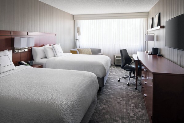 A hotel room with two beds, a desk, a chair, a TV, and a large window with blinds.