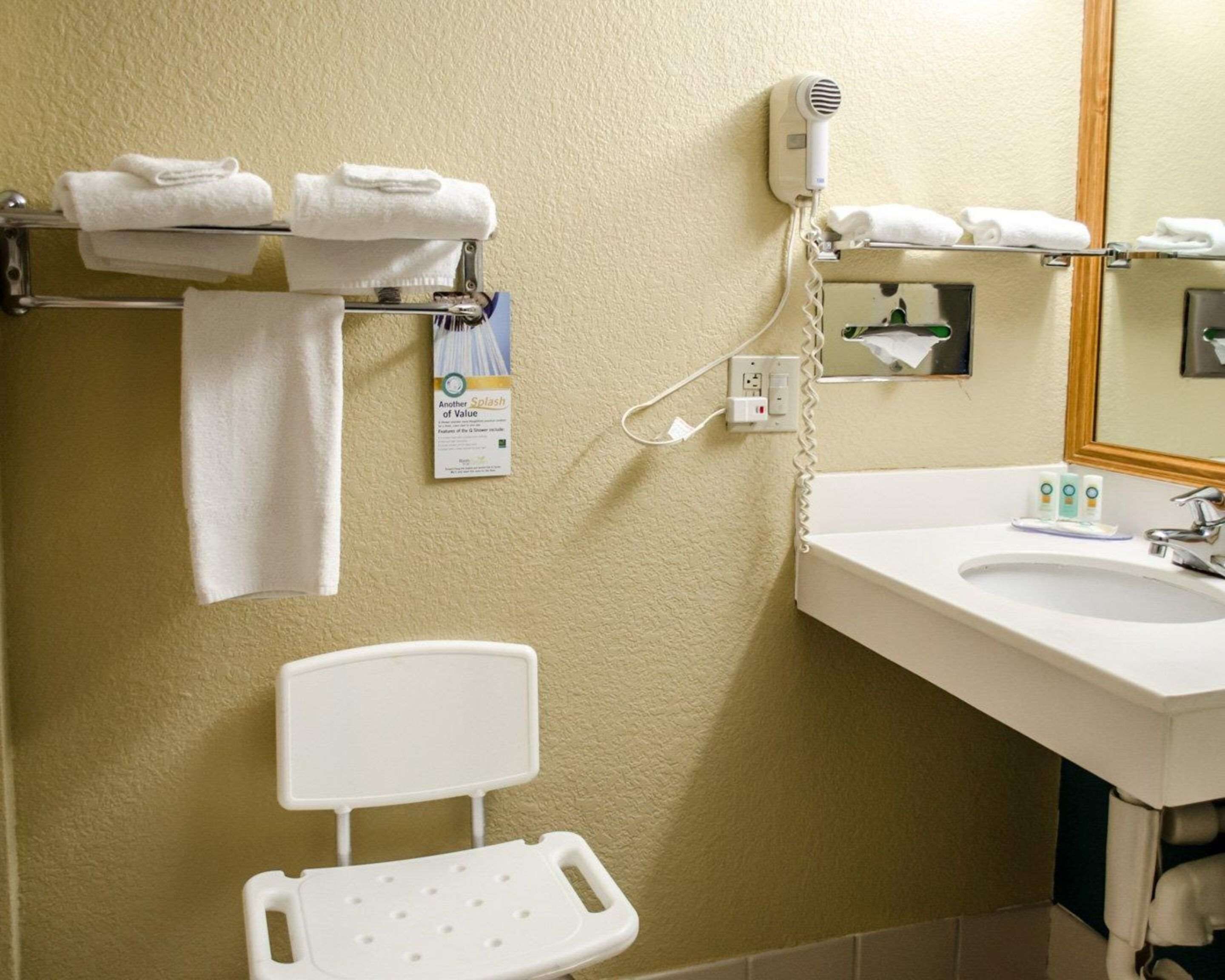 A bathroom with a sink, a mirror, a towel rack with folded towels, a portable shower chair, and a hairdryer plugged into a wall outlet.