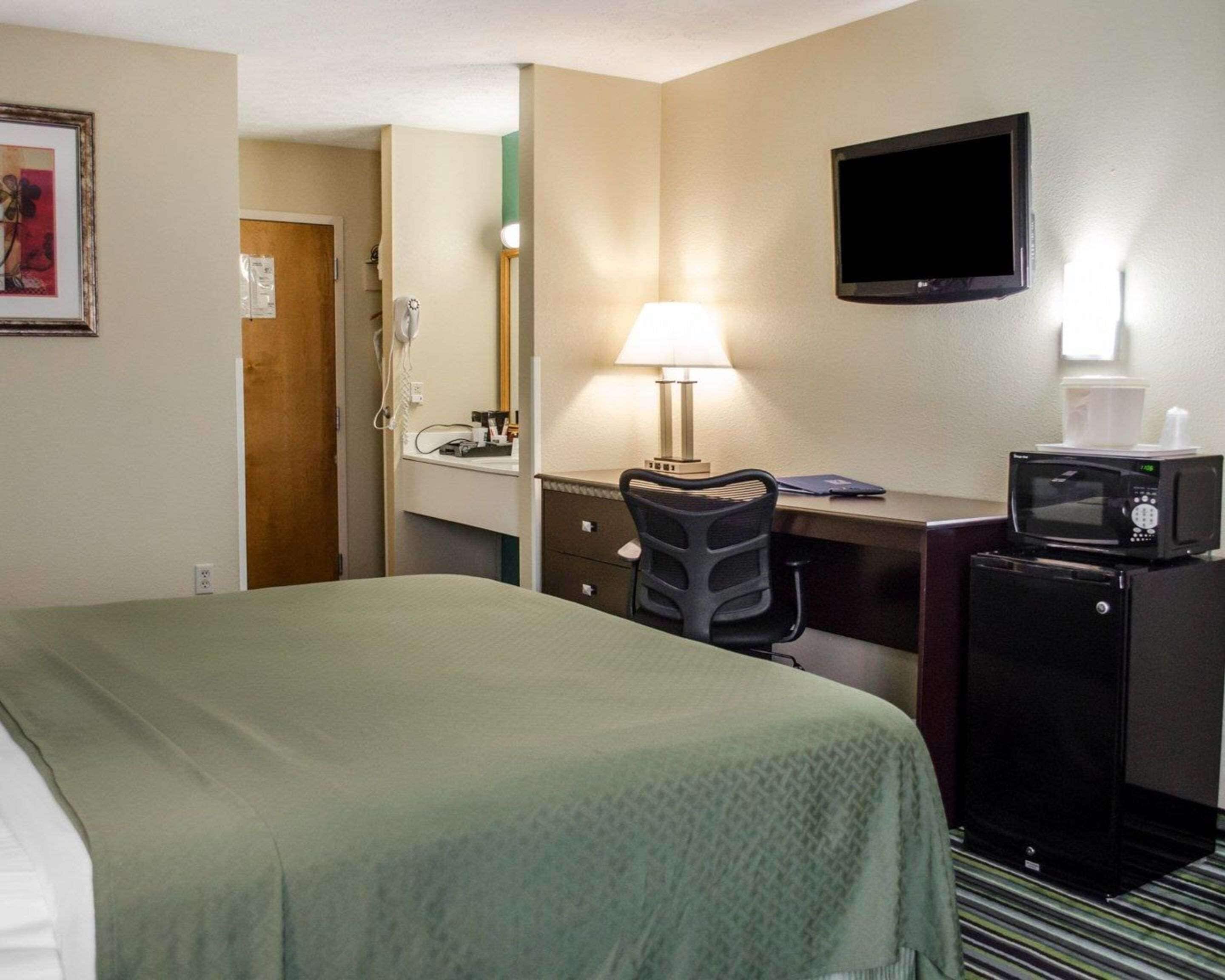 A hotel room with a bed, desk, chair, microwave, and a TV on the wall.