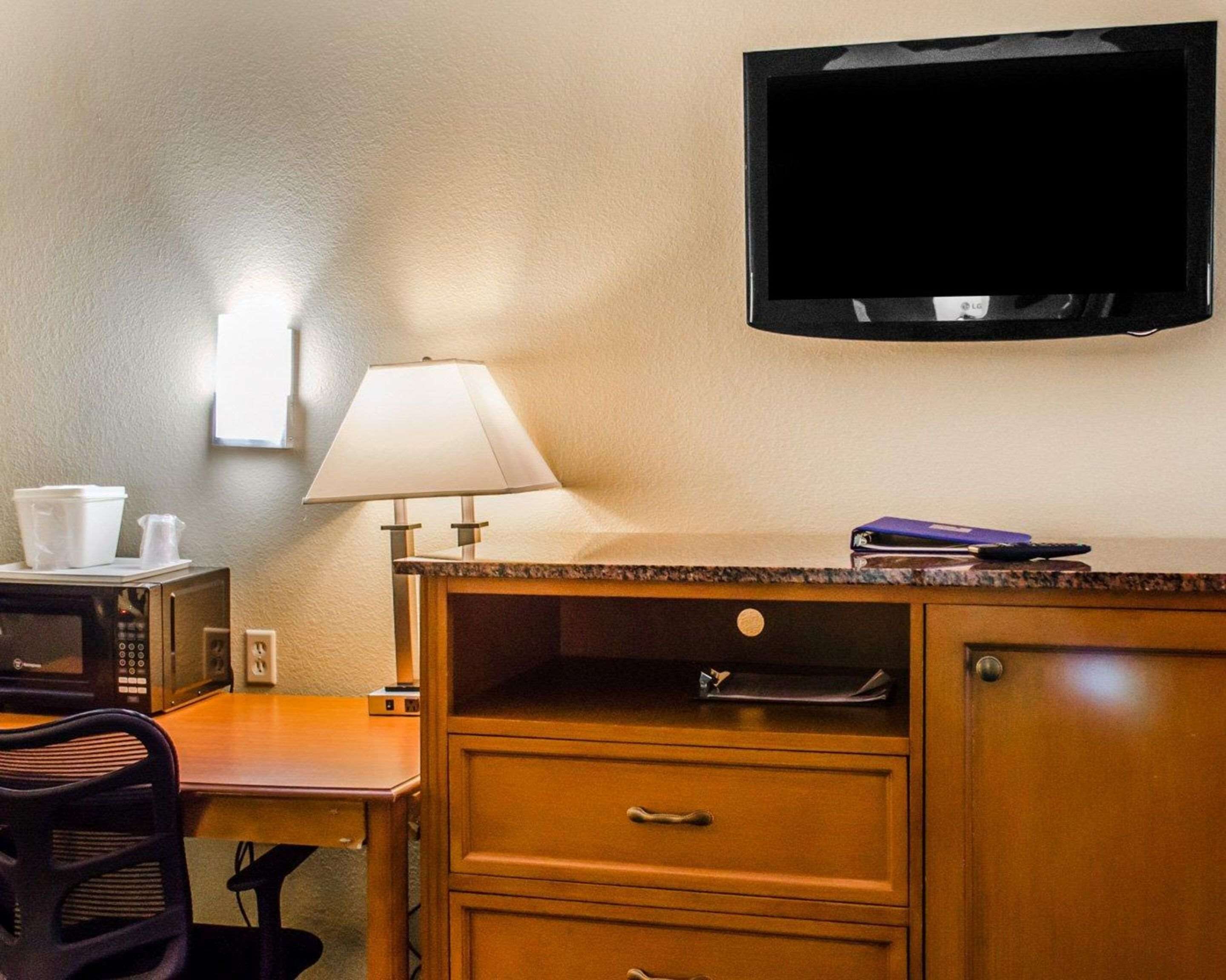 A hotel room with a wooden desk, a chair, a television mounted on the wall, a lamp, and a microwave on the counter.